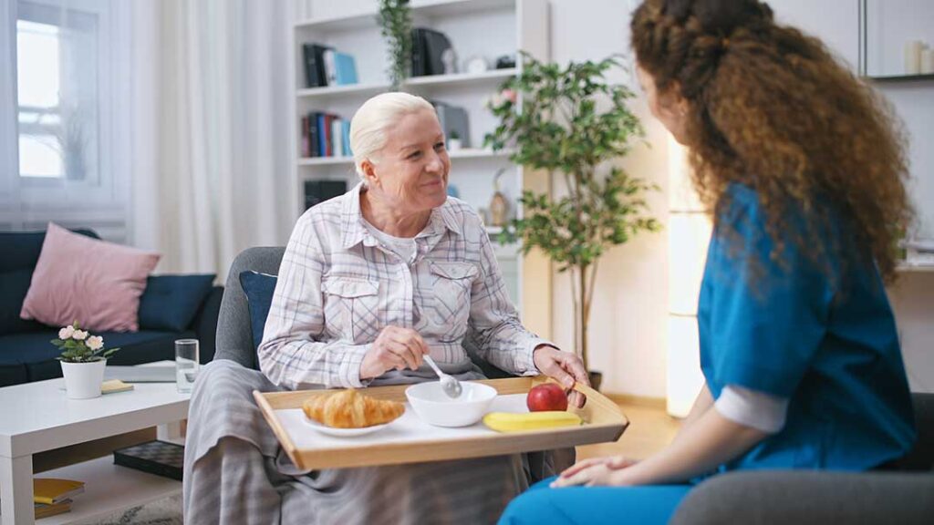 A senior resident sits in a cozy living room sharing her daily schedule with an attentive caregiver nearby.