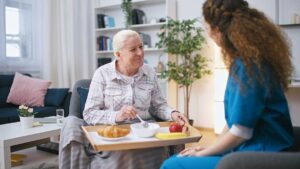 A senior resident sits in a cozy living room sharing her daily schedule with an attentive caregiver nearby.
