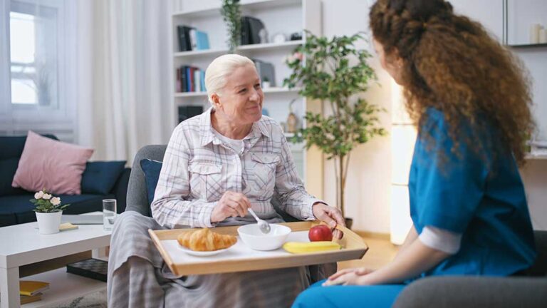 A senior resident sits in a cozy living room sharing her daily schedule with an attentive caregiver nearby.